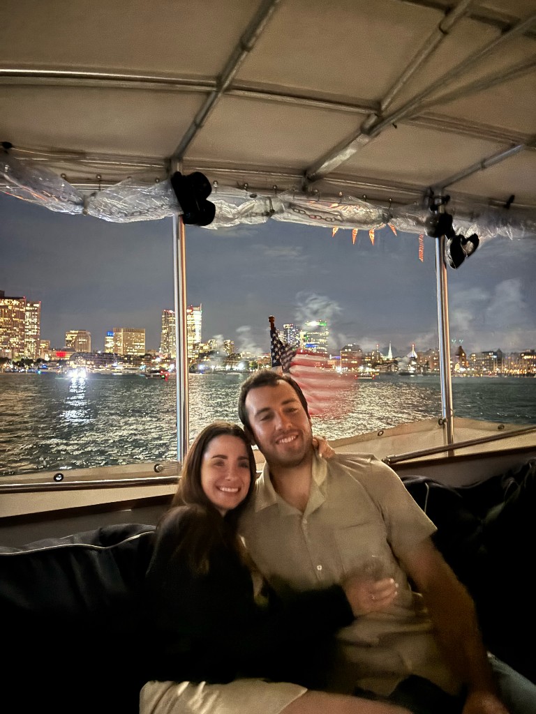 Jenna and Wyatt smiling together on a boat at night with city lights behind them